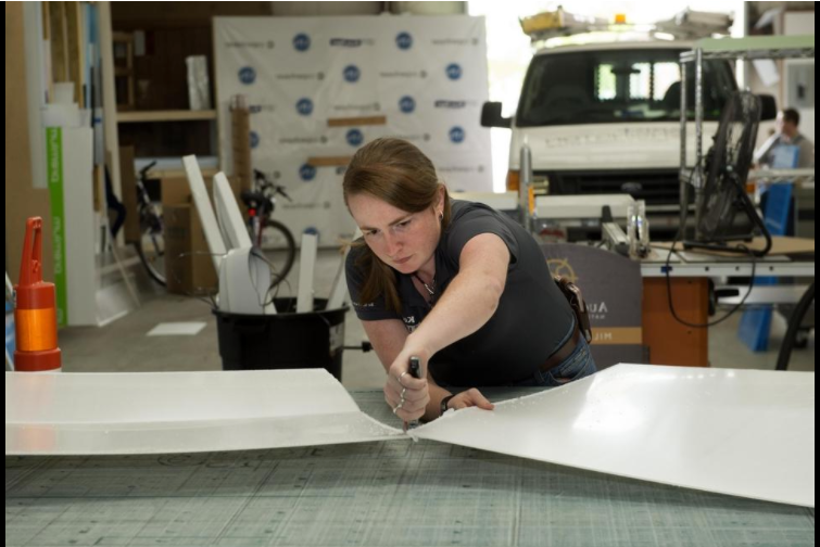 一头棕色头发的高加索女性, 布列塔尼·多斯特(Brittany Doster)斜靠在桌子上，正在切一块白板. 背景是一辆载着设计用品和工具的白色货车.