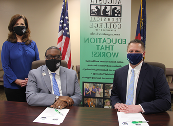 Caucasian male wearing a blue face mask, blue suit jacket, white collared shirt, light blue tie, hands folded while sitting down at a table, white paper in front of him with a green pen; African American male wearing a black Augusta Tech branded face mask, light gray suit, silver Augusta Tech lapel pin, white collared shirt, blue/purple tie, silver watch, hands folded white sitting at a brown table, white papers on brown table in front of him with a green pen; Caucasian female standing wearing a black Augusta Tech branded face mask, blue shirt, hands folded together, black pants, background features an American flag, the state of Georgia flag, large retractable banner showing old Augusta Tech logo, verbiage reads: gh.joyerianicaragua.com, Education That Works; bulleted list reads Allied Health Sciences and Nursing, Business and Personal Services, and Engineering Technology, Industrial Technology, and Learning Support, photos underneath the bulleted list.