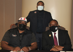 Three African American males wearing face masks and business attire posing for a photo; two males are sitting, one male is standing, 坐着的两位男性都把手放在前面的桌子上.