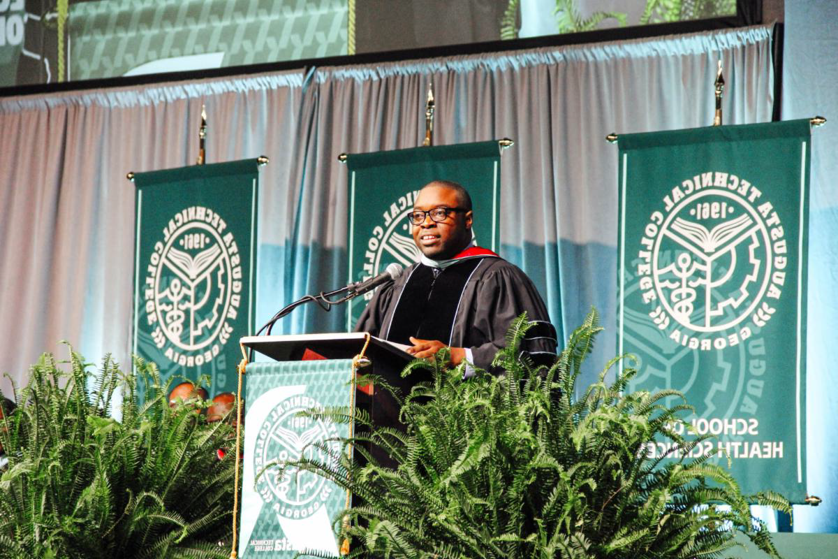 Dr. 身穿毕业典礼礼服的Whirl站在讲台上演讲，背景是学校的横幅，讲台的左右两侧是蕨类植物.